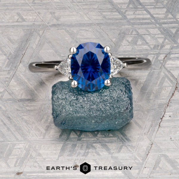The "Vesta" ring in platinum with 2.44-carat Montana sapphire The "Vesta" ring in platinum, set with a 2.44-carat Montana sapphire, is shown on a rough blue stone against a geometric metallic background featuring the "EARTH'S TREASURY" logo at the bottom.