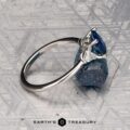 The "Vesta" ring in platinum, set with a 2.44-carat Montana sapphire, is shown on a rough blue stone against a geometric metallic background featuring the "EARTH'S TREASURY" logo at the bottom.