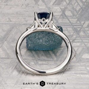 The "Vesta" ring in platinum, set with a 2.44-carat Montana sapphire, is shown on a rough blue stone against a geometric metallic background featuring the "EARTH'S TREASURY" logo at the bottom.