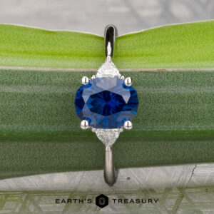 The "Vesta" ring in platinum, featuring a 2.44-carat Montana sapphire and two side stones, rests on a green leaf. The "Earth's Treasury" logo appears at the bottom.