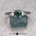 The Cathedral "Merope" ring in platinum features a 1.36-carat Australian sapphire center stone flanked by two pear-shaped clear stones, shown atop a rough blue crystal with the Earth’s Treasury logo below.