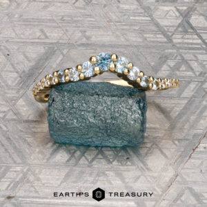 The "Venus" band in 18K yellow gold, featuring a row of white gemstones, is displayed on a rough blue-green stone atop a patterned gray surface, with the "Earth's Treasury" logo at the bottom.
