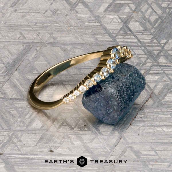 The "Venus" band in 18K yellow gold The "Venus" band in 18K yellow gold, featuring a row of white gemstones, is displayed on a rough blue-green stone atop a patterned gray surface, with the "Earth's Treasury" logo at the bottom.