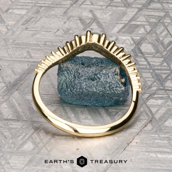 The "Venus" band in 18K yellow gold The "Venus" band in 18K yellow gold, featuring a row of white gemstones, is displayed on a rough blue-green stone atop a patterned gray surface, with the "Earth's Treasury" logo at the bottom.