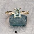 The "Waltz" ring in 18K yellow gold, featuring a 1.67-carat rectangular Montana sapphire and diamond accents, is displayed on a rough blue stone with the "Earth's Treasury" logo beneath.