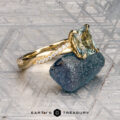 The "Waltz" ring in 18K yellow gold, featuring a 1.67-carat rectangular Montana sapphire and diamond accents, is displayed on a rough blue stone with the "Earth's Treasury" logo beneath.
