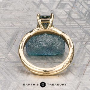 The "Waltz" ring in 18K yellow gold, featuring a 1.67-carat rectangular Montana sapphire and diamond accents, is displayed on a rough blue stone with the "Earth's Treasury" logo beneath.