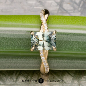 The "Waltz" ring in 18K yellow gold features a 1.67-carat square Montana sapphire center and diamond accents on the band, displayed on green with The Earth's Treasury logo elegantly at the bottom.