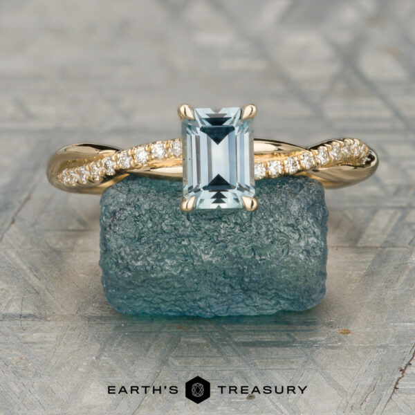 The "Waltz" ring in 14K yellow gold with 0.82-carat Montana sapphire The "Hambrick, Jesse and Burdue, Abigail 1" ring features a gold band with a rectangular aquamarine center stone and diamond accents, resting on a blue-green stone with the "Earth's Treasury" logo—a timeless piece for every moment.
