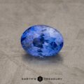 A close-up image of a 1.52-Carat Violet-Blue Ceylon Sapphire gemstone. The background is a textured, stone-like surface in a neutral color. The text “EARTH’S TREASURY” is visible at the bottom of the image.