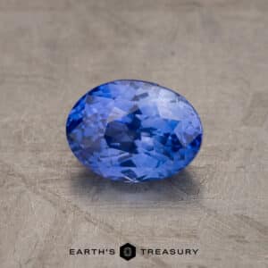 A close-up image of a 1.52-Carat Violet-Blue Ceylon Sapphire gemstone. The background is a textured, stone-like surface in a neutral color. The text “EARTH’S TREASURY” is visible at the bottom of the image.