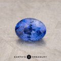 A close-up image of a 1.52-Carat Violet-Blue Ceylon Sapphire gemstone. The background is a textured, stone-like surface in a neutral color. The text “EARTH’S TREASURY” is visible at the bottom of the image.