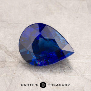 JM1023L features a pear-shaped, blue gemstone with intricate facets set against a textured gray surface, with the text "Earth's Treasury" displayed at the bottom.