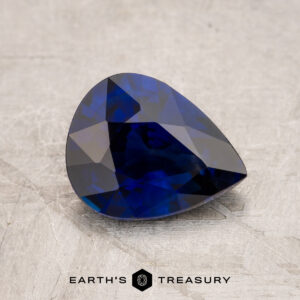 A faceted blue JM1026L gemstone on a textured surface with the "Earth's Treasury" logo at the bottom.