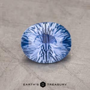 A 1.46-Carat Sky Blue Ceylon Sapphire sits on a textured, light gray surface. The intricate faceting creates a starburst pattern of light reflections within the stone. The name "EARTH'S TREASURY" is printed in black text below the gemstone.