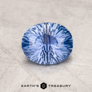 A 1.46-Carat Sky Blue Ceylon Sapphire sits on a textured, light gray surface. The intricate faceting creates a starburst pattern of light reflections within the stone. The name "EARTH'S TREASURY" is printed in black text below the gemstone.