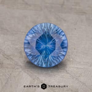 A round, faceted 1.69-carat medium blue Ceylon sapphire with intricate cut patterns rests on a textured gray surface. Below the gemstone, the text "Earth's Treasury" is visible, along with a hexagonal logo.