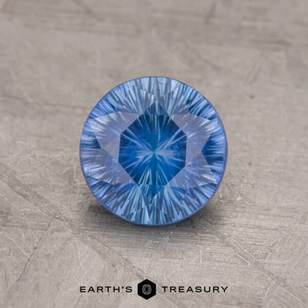 1.69-Carat Celyon Sapphire (Heated) A round, faceted 1.69-carat medium blue Ceylon sapphire with intricate cut patterns rests on a textured gray surface. Below the gemstone, the text "Earth's Treasury" is visible, along with a hexagonal logo.