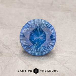 A round, faceted 1.69-carat medium blue Ceylon sapphire with intricate cut patterns rests on a textured gray surface. Below the gemstone, the text "Earth's Treasury" is visible, along with a hexagonal logo.