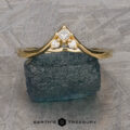 The "Sappho" Band, a gold ring featuring a triangular arrangement of three small diamonds above a larger central diamond, is displayed on top of a greenish-blue metallic rock. The setting has an elegant, pointed design. The text "Earth's Treasury" is visible at the bottom.