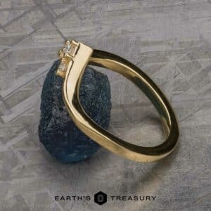 The "Sappho" Band, a gold ring with a minimalist design featuring two small square-cut diamonds, is elegantly displayed leaning against a rough, dark blue gemstone. The background has a textured, metallic appearance. The brand name "EARTH'S TREASURY" is visible at the bottom.