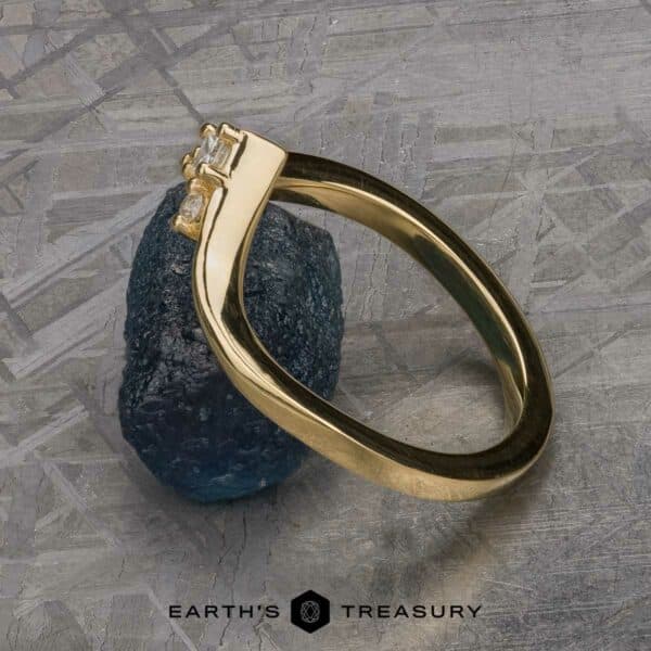 The "Sappho" band in 18k yellow gold The "Sappho" Band, a gold ring with a minimalist design featuring two small square-cut diamonds, is elegantly displayed leaning against a rough, dark blue gemstone. The background has a textured, metallic appearance. The brand name "EARTH'S TREASURY" is visible at the bottom.