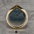 A close-up view of The "Sappho" Band, featuring a gold ring adorned with a teal-colored gemstone, captured from the side to highlight its intricate design. Set against a textured gray surface, the image includes the text "Earth's Treasury" at the bottom next to a small hexagon icon.