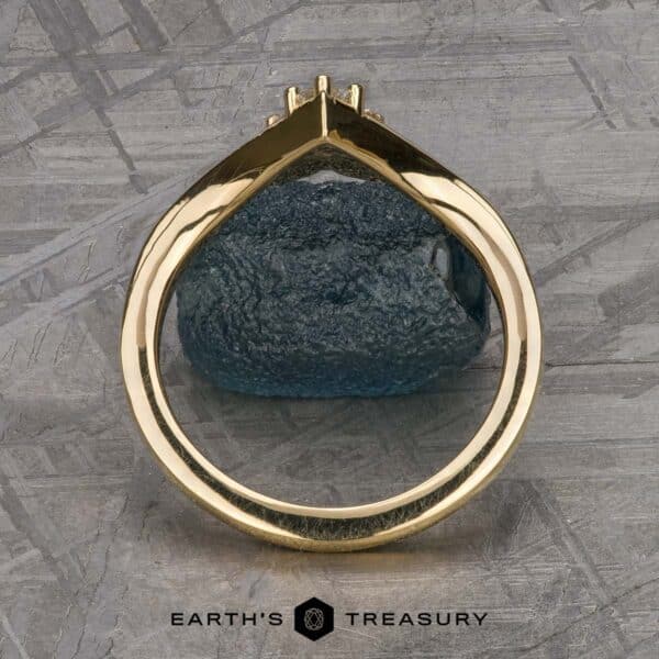 The "Sappho" band in 18k yellow gold A close-up view of The "Sappho" Band, featuring a gold ring adorned with a teal-colored gemstone, captured from the side to highlight its intricate design. Set against a textured gray surface, the image includes the text "Earth's Treasury" at the bottom next to a small hexagon icon.