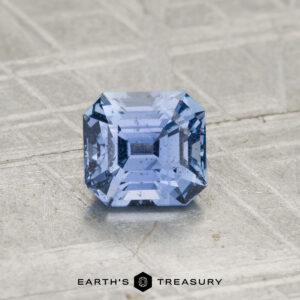 The 1.26-Carat Violet-Blue Sapphire, square-cut with visible inclusions, rests on a textured light gray surface. "EARTH'S TREASURY" appears at the bottom.