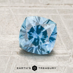 The MS7944, a square brilliant-cut Montana Sapphire, rests on a textured light surface with the Earth’s Treasury logo displayed underneath.