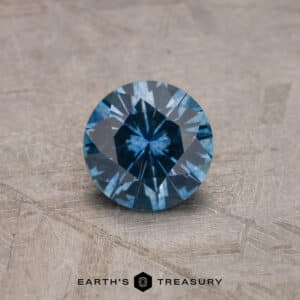 A close-up image of a 2.14-carat Robin's Egg Blue Montana Sapphire with intricate facets, positioned on a textured gray surface. The gemstone has a deep, vivid blue color. At the bottom center of the image is the text "Earth's Treasury" accompanied by a small logo.