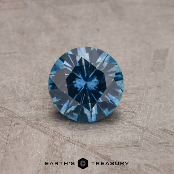 1.17-Carat Montana Sapphire (Heated) A close-up image of a 2.14-carat Robin's Egg Blue Montana Sapphire with intricate facets, positioned on a textured gray surface. The gemstone has a deep, vivid blue color. At the bottom center of the image is the text "Earth's Treasury" accompanied by a small logo.