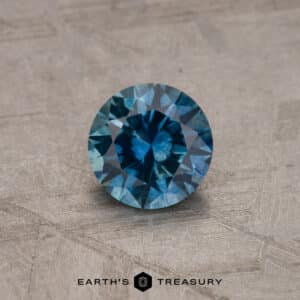 A close-up of a round, robin's egg blue Montana sapphire with intricate facets. It is placed on a textured, light brown surface. The bottom of the image features the text "EARTH'S TREASURY" accompanied by a geometric logo.