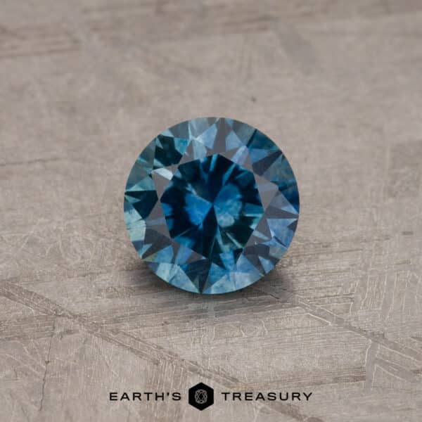 1.17-Carat Montana Sapphire (Heated) A close-up of a round, robin's egg blue Montana sapphire with intricate facets. It is placed on a textured, light brown surface. The bottom of the image features the text "EARTH'S TREASURY" accompanied by a geometric logo.