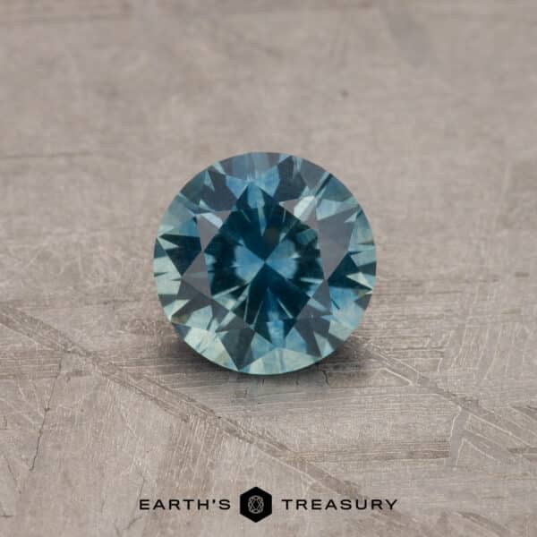 1.14-Carat Montana Sapphire (Heated) A 2.14-carat robin's egg blue Montana sapphire (heated) is displayed on a textured gray surface. The gemstone has a brilliant cut, showcasing various facets that reflect light beautifully. Below the gemstone, the text "Earth's Treasury" is visible.