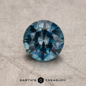 A vivid blue, round faceted 1.05-carat heated Montana Sapphire is shown on a textured surface with the "Earth's Treasury" logo and text at the bottom.