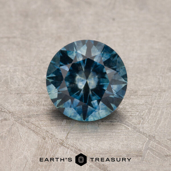 1.05-Carat Montana Sapphire (Heated) A vivid blue, round faceted 1.05-carat heated Montana Sapphire is shown on a textured surface with the "Earth's Treasury" logo and text at the bottom.
