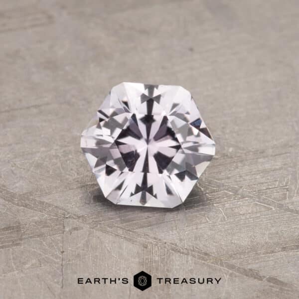 0.91-Carat Montana Sapphire A faceted hexagonal 0.91-carat near colorless Montana sapphire with a brilliant cut, displaying clear and sparkling clarity, is placed on a textured gray surface. The text "EARTH'S TREASURY" is centered below the gemstone.