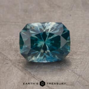 A close-up image of a faceted 2.14-carat Robin's Egg Blue Montana sapphire gemstone resting against a textured grey background. The complex, symmetrical cuts of the gem create a sparkling effect. At the bottom center, the logo and text "EARTH'S TREASURY" is visible.
