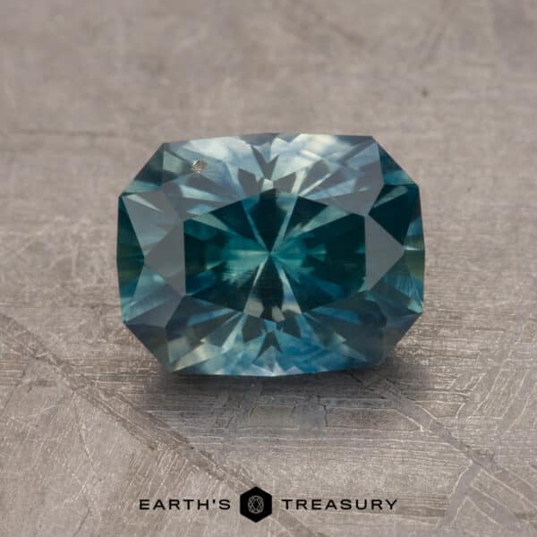1.68-Carat Montana Sapphire (Heated) A close-up image of a faceted 2.14-carat Robin's Egg Blue Montana sapphire gemstone resting against a textured grey background. The complex, symmetrical cuts of the gem create a sparkling effect. At the bottom center, the logo and text "EARTH'S TREASURY" is visible.