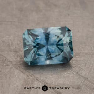 A close-up image of a rectangular, faceted blue-green gemstone placed on a textured gray surface. The stone has a symmetrical cut with multiple facets reflecting light. The text "2.14-Carat Robin's Egg Blue Montana Sapphire (Heated)" is visible at the bottom of the image.