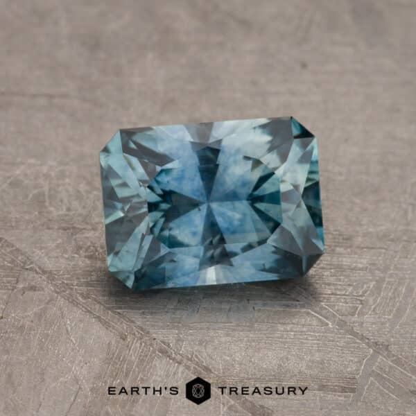 1.27-Carat Montana Sapphire (Heated) A close-up image of a rectangular, faceted blue-green gemstone placed on a textured gray surface. The stone has a symmetrical cut with multiple facets reflecting light. The text "2.14-Carat Robin's Egg Blue Montana Sapphire (Heated)" is visible at the bottom of the image.