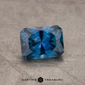 A 2.14-Carat Robin's Egg Blue Montana Sapphire (Heated) with a rectangular cut and intricate facets is displayed on a textured surface. The gemstone showcases a rich color and sparkling inclusions. At the bottom, the image features the logo and text "Earth's Treasury.