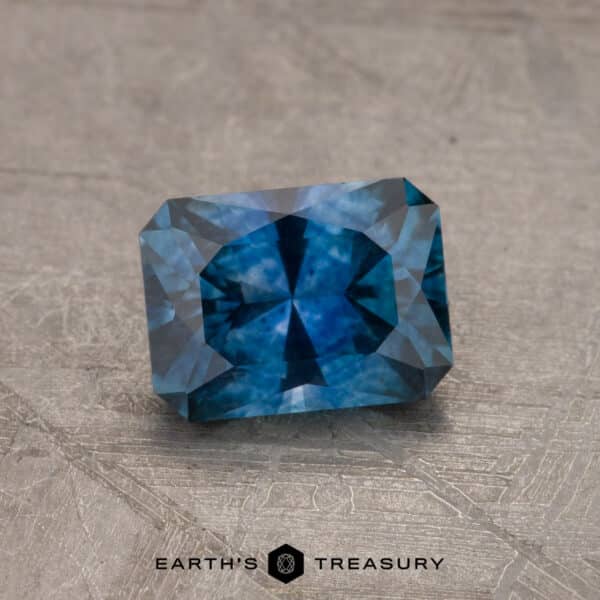 1.14-Carat Montana Sapphire (Heated) A 2.14-Carat Robin's Egg Blue Montana Sapphire (Heated) with a rectangular cut and intricate facets is displayed on a textured surface. The gemstone showcases a rich color and sparkling inclusions. At the bottom, the image features the logo and text "Earth's Treasury.