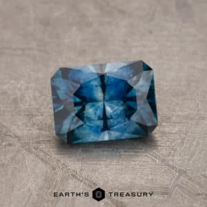 A 2.14-carat Robin's Egg Blue Montana Sapphire, expertly heated to enhance its vivid shades of blue, with intricate facets, rests on a textured gray surface. The gemstone reflects light strikingly and appears impeccably crafted and polished. At the bottom of the image, "EARTH'S TREASURY" is prominently displayed.