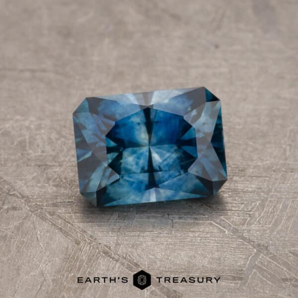 1.08-Carat Montana Sapphire (Heated) A 2.14-carat Robin's Egg Blue Montana Sapphire, expertly heated to enhance its vivid shades of blue, with intricate facets, rests on a textured gray surface. The gemstone reflects light strikingly and appears impeccably crafted and polished. At the bottom of the image, "EARTH'S TREASURY" is prominently displayed.
