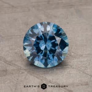 An intricately cut, round Robin's Egg Blue Montana Sapphire gemstone with a brilliant 2.14-carat sparkle rests on a textured, stone-like surface. At the bottom of the image, the text "EARTH'S TREASURY" is displayed.