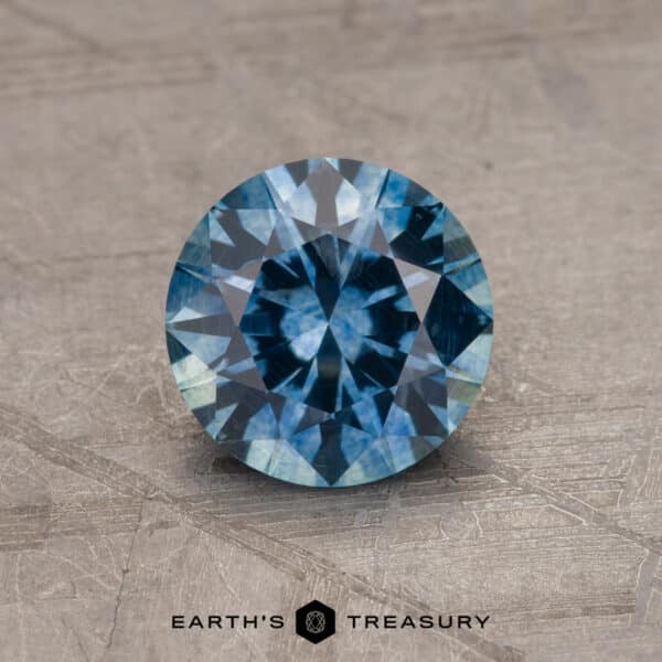 1.69-Carat Montana Sapphire (Heated) An intricately cut, round Robin's Egg Blue Montana Sapphire gemstone with a brilliant 2.14-carat sparkle rests on a textured, stone-like surface. At the bottom of the image, the text "EARTH'S TREASURY" is displayed.