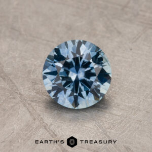 A 1.44-carat sky blue Montana Sapphire (heated), round brilliant-cut, is shown on a textured surface with the "Earth's Treasury" logo below it.
