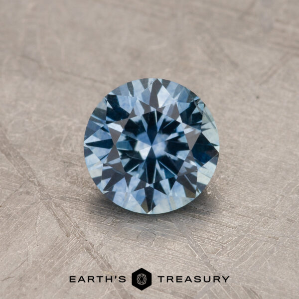 1.44-Carat Sky Blue Montana Sapphire (Heated) A 1.44-carat sky blue Montana Sapphire (heated), round brilliant-cut, is shown on a textured surface with the "Earth's Treasury" logo below it.